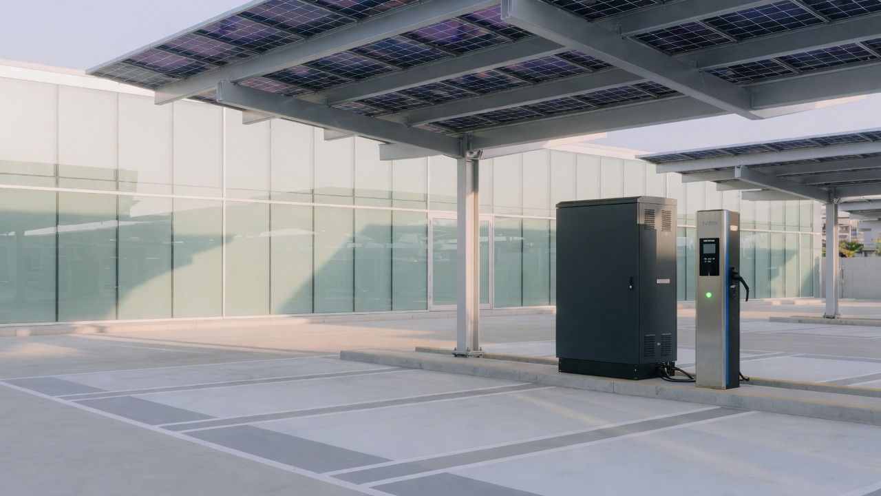 Estacionamento coberto por placas solares, com um gabinete de baterias ao lado de um carregador.